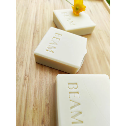 Three earth yellow clay soap bars laid on a wooden board with a small yellow poppy.  The soaps are stamped with BEAM. 