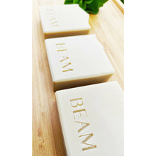 Load image into Gallery viewer, Three earth yellow clay soap bars on a wooden board with some green foliage.  The soaps are stamped with BEAM. 
