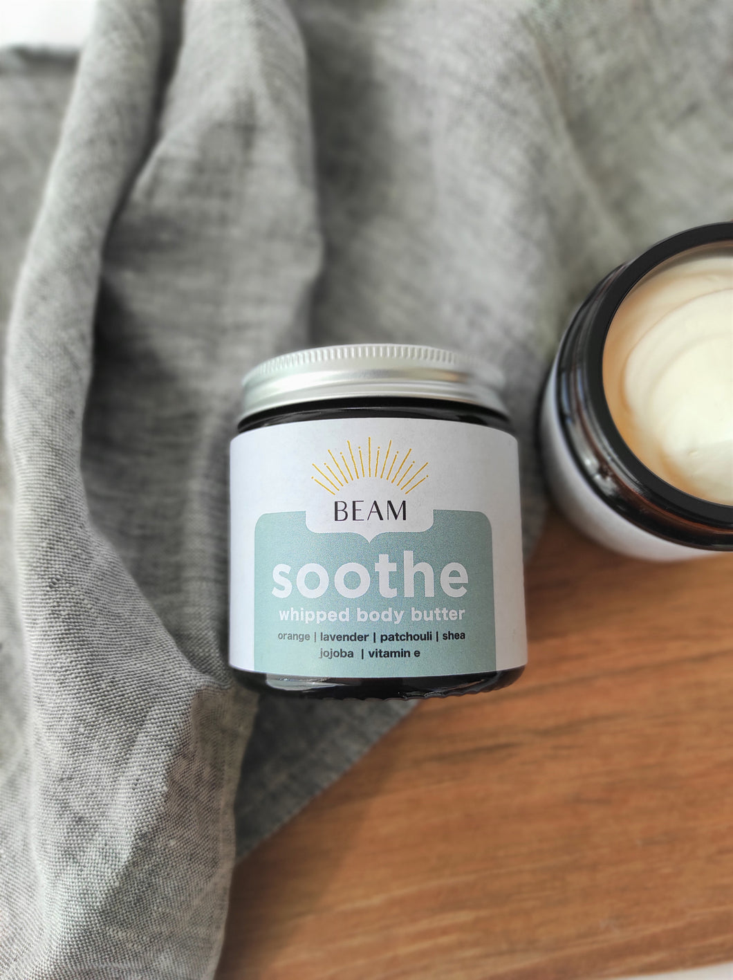 An amber glass jar of whipped body butter with silver metal lid.  The label is white and blue with soothe  printed on it and the BEAM logo.